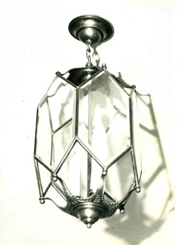 Lampa mosiężna, trzy świecowa z kryształowymi szkłami, sześcioboczna w typie latarni XIX wiecznej - wisząca. Wokół mosiężnego pręta, stanowiącego trzon konstrukcji rozmieszczono trzy stylizowane świece z żarówkami. Do pręta od góry i od dołu przymocowano mosiężne dekoracyjne formy zbliżone do owalu, z których każdy rozwidla się na sześć ozdobnych łapek, połączonych kulkami z mosiężną konstrukcją sześciobocznego kryształowego przeszklenia lampy.